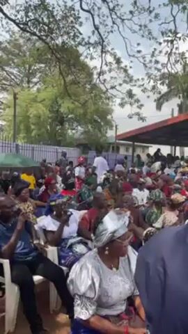The video shows a large gathering of people, primarily adults, seated in rows of white plastic chairs, with some under a sheltered structure with a red roof. The crowd is diverse in age and attire, with many wearing traditional African clothing. Some individuals are using phones, while others are listening to a speaker who is not visible in the frame. Trees surround the area, suggesting an outdoor setting. The lighting appears natural, indicating a daytime event. Overall, the scene depicts a formal gathering or community event taking place outdoors.