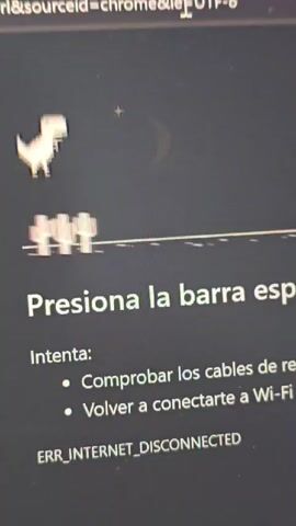 A close-up shot shows a computer screen displaying the Google Chrome dinosaur game, which appears when the internet is disconnected. The white dinosaur is running along a line, jumping over cacti. Text on the screen reads "Presiona la barra esp". Below that, two bullet points read "Comprobar los cables de re" and "Volver a conectarte a Wi-Fi." The bottom of the screen reads "ERR_INTERNET_DISCONNECTED".  The screen is somewhat dirty.