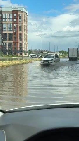The video shows a white car driving through a flooded street. The car passes in front of the camera, splashing water as it moves. The scene is shot from inside another car, looking through the windshield. The water level is high, covering the road entirely. Buildings are visible in the background. The weather is overcast with some blue sky. The flooded street is surrounded by sidewalks and greenery. Other cars and trucks are visible in the distance on the higher ground.