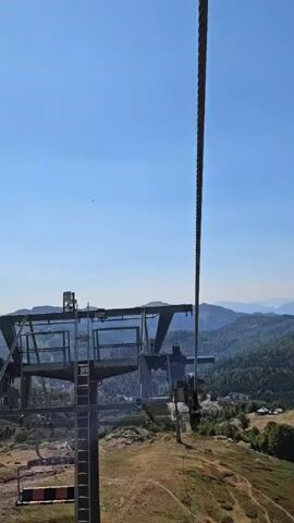 The video shows a close-up view of a ski lift mechanism against a clear blue sky. The ski lift tower has metal platforms and stairs, with a thick cable running through it. The cable is the main focus, as it extends upwards and out of frame. The background shows a mountainous landscape with green trees and some scattered buildings. The sun is visible in the upper left corner. The camera angle is looking upwards, from a lower position. The ski lift appears to be stationary.