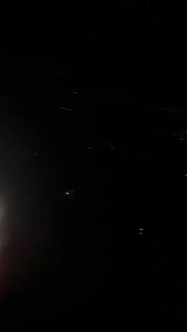 The video shows a night view from an airplane window. The scene below is dark with scattered lights, indicating a city or town. The lights appear as small white or yellow dots, some forming patterns that suggest streets or buildings. The perspective is from a high altitude, looking down at the landscape. The camera is slightly shaky, suggesting movement, likely from the airplane in flight. The top of the frame is completely black, representing the night sky. The bottom right corner shows a sliver of the airplane's wing or fuselage.