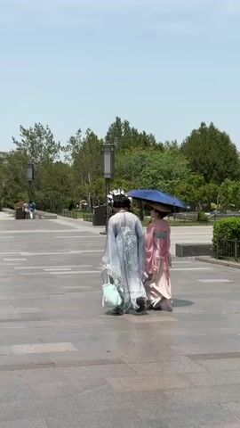 Two women in traditional Chinese clothing are walking away from the camera on a wide, paved walkway. The woman on the left is wearing a light blue robe with floral patterns and has a large floral headpiece. She carries a light blue bag. The woman on the right is wearing a pink and cream-colored robe and holds a blue umbrella over herself. The walkway is lined with trees, lamp posts, and other people walking in both directions. In the background, there are traditional Chinese-style buildings. The sky is blue and clear.