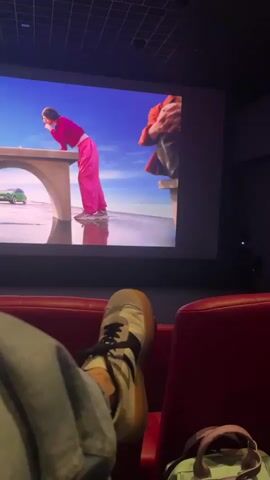Here is a concise description of the video:

The video shows a person sitting in a red movie theater seat, with their legs crossed and one foot visible. They are watching a large movie screen. The screen displays scenes from a brightly colored film. The film includes a woman in pink overalls, a man in a red-toned outfit, an aerial view of a pink-toned race track, and close-ups of a green car. The theater is dimly lit. A light green bag is placed on the seat next to the person.