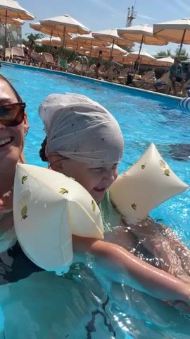 A woman in sunglasses and a dark swimsuit smiles at the camera, holding a child in a swimming pool. The child wears a light-colored headscarf and arm floaties. Water splashes around them. In the background, lounge chairs, umbrellas, and other people are visible near the pool.