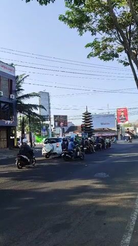 The video shows a street scene with several motorbikes lined up, waiting at what appears to be an intersection or traffic signal. A white car is positioned in front of the motorbikes. Buildings line the left side of the street, including a multi-story building with signage. Trees with green foliage are visible, and power lines crisscross the sky. A pagoda-like structure is present in the background, along with billboards. The lighting suggests it is daytime, possibly late morning or early afternoon. The overall atmosphere is one of typical street activity.