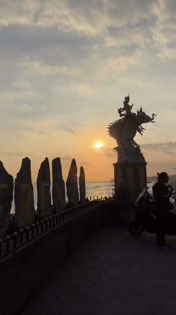 Sunset gathering at Indonesian beach features mythological statues, kite flying