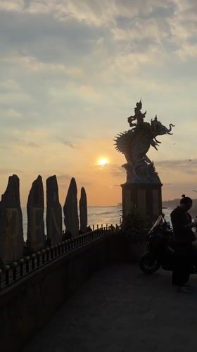 Sunset gathering at Indonesian beach features mythological statues, kite flying