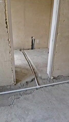 The video shows an interior scene with concrete walls and floor. A doorway is visible with exposed foam insulation around the frame.  White pipes are laid on the floor, and one pipe is partially buried in a trench. In the background, a hole is visible in the wall with pipes protruding from it, and a jackhammer is leaning against the wall. The scene appears to be under construction or renovation.