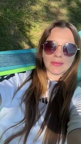A woman with long brown hair and sunglasses lies in a hammock outdoors. She is wearing a white t-shirt. The hammock has blue, green, and white stripes. Sunlight filters through the trees, casting shadows on her face and shirt.