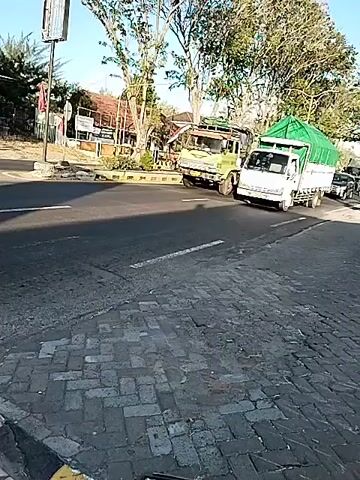 The video shows a street scene with several vehicles and pedestrians. In the foreground, there is a cobbled surface and a road with white lines. A white truck with a green tarp covering its cargo is in the middle ground, turning to the right. To its left, a green truck is moving in the opposite direction. A silver SUV and a dark-colored minivan are also on the road. Further in the background, there are trees, buildings, and other vehicles. Two pedestrians are visible in the foreground, one wearing a hijab and carrying a backpack, crossing the street. Another person, partially visible, is walking behind them. The lighting suggests it is daytime.