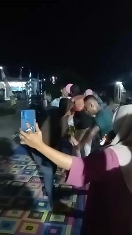 A nighttime scene shows a group of people gathered outdoors. In the foreground, a person wearing a headscarf and a purple shirt is holding a phone, seemingly recording the event. Behind them, several individuals are engaged in an activity, with some holding balloons. One person in a brown shirt is leaning forward, and another in a dark green shirt is in a crouched position. The ground is covered with a patterned mat. In the background, there are lights and structures suggesting a residential area. The overall atmosphere appears festive or celebratory.
