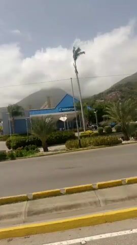 The video shows a street scene in Caraballeda, Venezuela, on a cloudy day. The camera pans from left to right, capturing a busy road with cars and a motorcycle. In the background, lush green mountains rise up, dotted with houses. 

On the left side of the road, a building with a blue roof and white trim is visible. A tall, slender palm tree stands in front of it, its fronds swaying in the breeze. A motorcycle with a rider wearing a white shirt and a helmet is seen cruising down the road in the middle of the frame. 

Further down the road, there are more trees and buildings, and the mountains continue to dominate the background. A few cars are parked along the roadside, and a person can be seen walking on the sidewalk in the distance. The overall impression is of a tropical city on a partly cloudy day, with vibrant green vegetation and a mix of modern and older architecture. There is no visible text in the video.