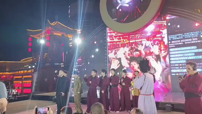 The video captures an outdoor evening event on a stage with a large LED screen in the background displaying moving images of people with their hands raised. A group of men dressed in traditional Chinese attire, primarily in maroon robes with some in black and tan suits, stand on the stage. One individual on the far right, also in maroon, holds a microphone and appears to be speaking or performing. Another person in a purple robe and a black hat stands to the left of the stage, also holding a microphone. The stage is set against a backdrop of traditional Chinese architecture illuminated with red lights. Several people in the foreground are holding up smartphones, presumably recording the event. The overall atmosphere suggests a performance or a public gathering.
