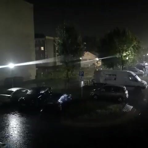 A night scene shows a street with several cars parked on the wet pavement. A white van is parked in the background. Trees are visible on the side of the street. Streetlights illuminate the scene, reflecting on the wet surfaces. It appears to be raining, as indicated by the wet ground and possible streaks in the air. The overall atmosphere is dark and wet.