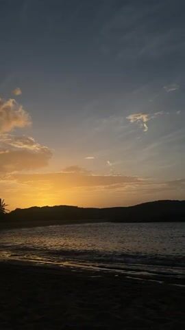 The video shows a sunset over a beach. The sky is a gradient of blue and orange, with some clouds. The sun is low on the horizon, casting a warm glow on the scene. The ocean is calm, with gentle waves lapping at the shore. The beach is dark, with the sand appearing black in the fading light. A person is walking along the beach in the distance. The overall mood is peaceful and serene.