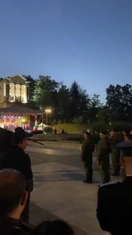 At dusk, a gathering of soldiers in camouflage uniforms and black berets stand at attention on a paved plaza. Behind them is a stage and a white building illuminated with lights. Several people in casual clothing are present. The camera angle is slightly low, looking up at the soldiers. A few soldiers with shields and rifles move forward. Music plays in the background.