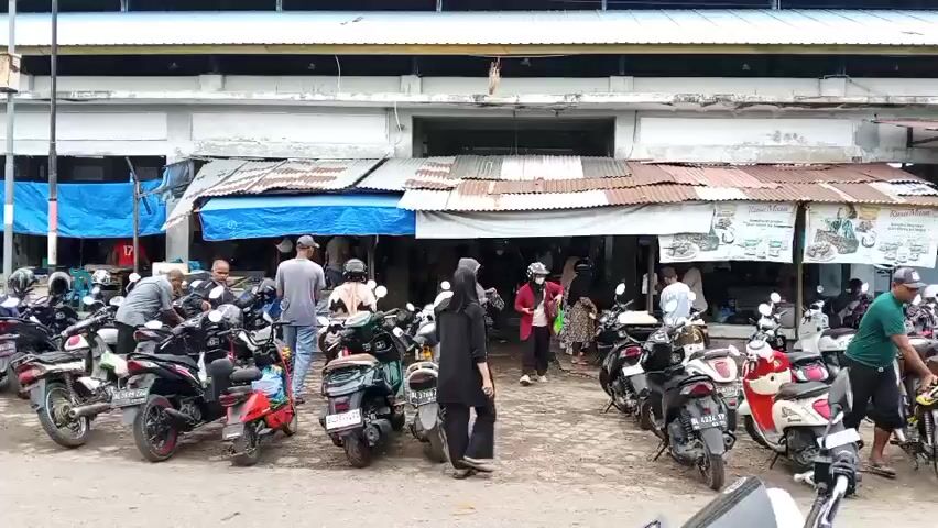 The video shows a busy marketplace scene with a large number of parked motorcycles. In the background, there is a long building with awnings and stalls, suggesting it is an indoor/outdoor market. The building has a sign that appears to read "PASAR IKAN", which translates to "Fish Market". People are seen walking around, some are riding motorcycles, and others are interacting near the stalls. The ground is paved with rough stones or gravel. The overall atmosphere appears to be one of commerce and daily activity in a market setting.