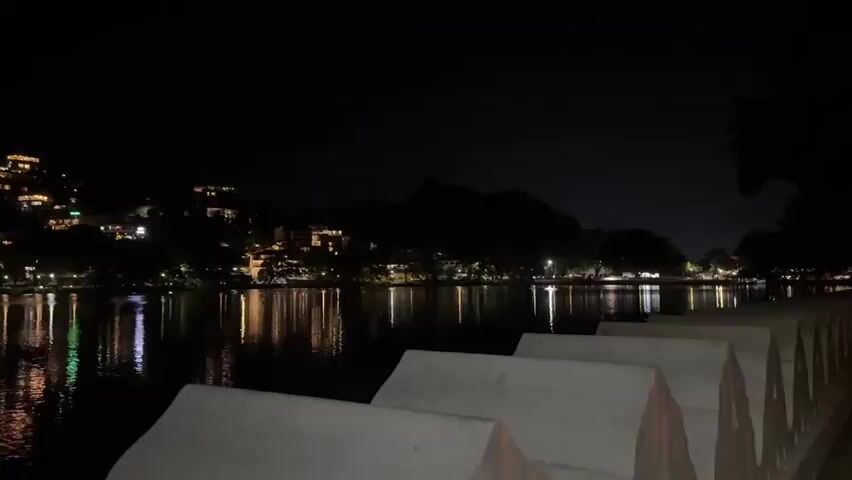 The video shows a nighttime view of a body of water, likely a lake, with lights reflected on the surface.  On the opposite shore, there are buildings and structures illuminated, scattered across a dark hillside. The foreground includes a portion of a light-colored, possibly concrete, barrier. The sky is dark, suggesting a clear night.  The lighting comes from the buildings and other light sources along the shoreline. There are no people visible in the video.