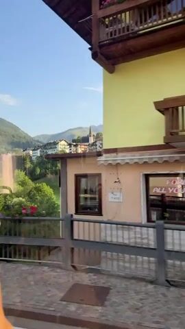 A video captures a street scene in a European town. The buildings are colorful, with balconies adorned with flowers. A restaurant with a sign that says "FOREVER ALL YOU CAN EAT" is visible. There are street signs and cobblestone streets. The video appears to be shot from a moving vehicle.