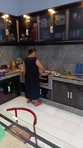 A woman in a dark dress stands in a kitchen, facing a stove. She is using a utensil to stir something in a pan on the stove. The kitchen has dark cabinets with glass doors and light-colored countertops. There is a tiled floor and a table with a red chair in the foreground.