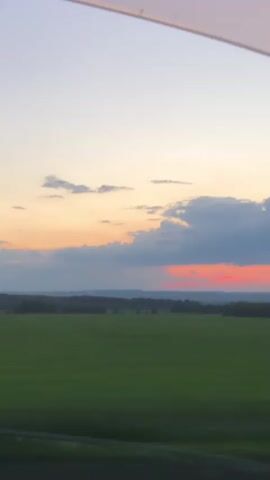 A video captures a scenic view from a vehicle, likely a train, as it travels through a rural landscape in Berezovyi, Russia. The camera is positioned to look out of a window, with the top edge of the window frame visible at the top of the screen.

The foreground is dominated by a vast, lush green field, suggesting farmland or pasture. Beyond the field, a dense line of dark green trees forms a horizon, interspersed with rolling hills in the distance. The sky above is a canvas of a sunset, displaying a gradient of colors. The upper portion of the sky is a pale blue, transitioning into soft oranges and yellows near the horizon. Fluffy white and grey clouds are scattered across the sky, with a particularly dramatic bank of darker clouds to the right, illuminated by the setting sun with a vibrant streak of pink and orange.

The overall mood is tranquil and serene, evoking the peacefulness of a rural evening. The soft colors of the sunset create a warm and calming atmosphere. The time of day is clearly evening, as indicated by the sunset. There is no visible text in the video. The activity taking place is the passive observation of the landscape from a moving vehicle.