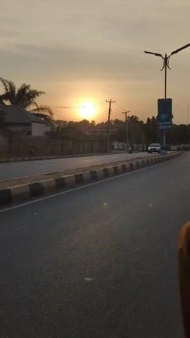 The video shows a sunrise scene on a road. The sun is low on the horizon, obscured by trees and power lines. A road runs along the bottom of the frame with a black and white curb. A sign and streetlight stand on the right side of the road. A few pedestrians walk along the side of the road. Vehicles, including a car, a motorcycle, and a yellow auto rickshaw labeled "NARAYI BUS STOP 6", pass by.