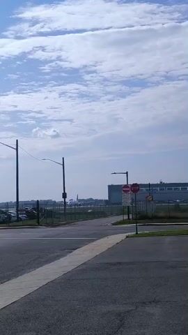 The video shows an outdoor scene with a bright blue sky filled with scattered clouds. In the foreground, there is a paved road with a concrete curb and a white pedestrian crossing. Beyond the road, a chain-link fence separates the area from a parking lot filled with cars. Further in the distance, an airplane with the Canadian flag on its tail is visible, appearing to be parked or taxiing on a runway. Two tall light poles stand on either side of the road, and a stop sign with additional regulatory signs is mounted on one of them. The overall environment suggests an airport setting on a clear day.