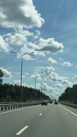 This is a first-person perspective video of a car driving on a highway. The road has multiple lanes and is surrounded by trees on either side. There are streetlights lining the highway. The sky is blue with fluffy white clouds. Other cars are visible on the road, including a few cars ahead and some in the oncoming lanes. The sun appears to be shining, illuminating the scene. There are no people visible outside of the vehicles. The overall environment is that of a typical highway in a wooded area on a sunny day.