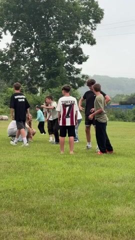 This video shows a group of young people in a grassy field. In the foreground, a person is sitting on the grass on the left side of the frame, and another person is also sitting down nearby. A third person is crouched down. Further to the right, a person in a white and red jersey with the number 7 is standing and facing away from the camera. To their right, two more people are standing and appear to be interacting. One is wearing a black t-shirt and shorts, and the other is wearing a green t-shirt and black pants. The background is filled with trees and a slightly overcast sky. The ground is covered in green grass.
