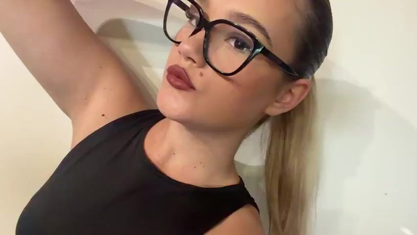 This media file features a close-up, waist-up shot of a young woman with light skin and blonde hair pulled back in a ponytail. She is wearing large, black-rimmed glasses and a black sleeveless top. Her makeup includes dark lipstick and what appears to be a mole or beauty mark on her cheek. Her nails are long and pointed, with some painted in bright colors like yellow and pink, and others with glitter.

The woman is leaning against a plain, off-white wall, and the lighting suggests an indoor setting. The overall mood is playful and flirty, as she is posing with her head tilted and her hand near her face, with her tongue visible in some frames. The scene is focused entirely on the woman, with no other objects or people present. The context of Beguntsi, Bulgaria, is provided but not visually evident within the frame. There is no visible text in the media file.