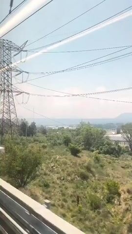 The video shows the view from a moving train looking out towards a landscape. The landscape consists of green vegetation, trees, and some buildings in the distance. The sky is light blue with some clouds. In the foreground, a large metal power line tower is visible with numerous power lines extending across the sky. A metal fence runs parallel to the train track. The video is shot from inside the train.