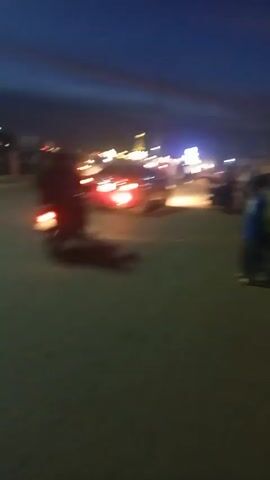 The video shows a street scene at dusk. The sky is a deep blue, transitioning to lighter shades near the horizon. Streetlights and vehicle headlights illuminate the scene. Several people are standing or walking on the paved road and sidewalks. A person on a motorcycle is driving away from the camera. In the distance, there appears to be a structure with multiple illuminated sections, suggesting a building or attraction. The video quality is somewhat blurry.