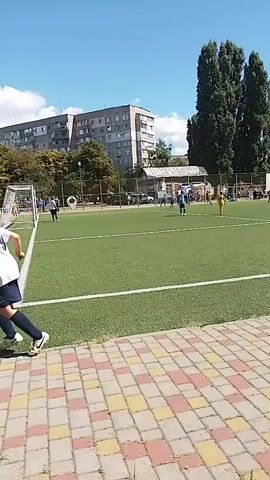 The video shows a boy in a white shirt and blue shorts playing soccer on an artificial turf field on a sunny day. He is running towards the right side of the frame with the ball at his feet. In the background, other players in blue jerseys are visible, as well as a soccer goal on the left side of the frame. There are trees, buildings, and a blue sky in the background. The foreground shows a checkered pattern of red, yellow, and beige paving stones.
