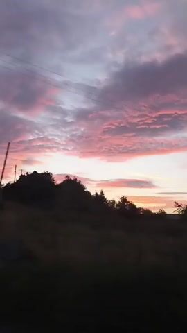 The video shows a time-lapse recording of a sunset over a rural landscape. The sky is filled with dramatic clouds in shades of purple, pink, and orange, indicating the sun is low on the horizon. In the foreground, silhouetted trees and a dark grassy field are visible. As the video progresses, a white house with a dark roof comes into view, situated among the trees. A power pole with electrical equipment is also visible. The overall atmosphere is serene and picturesque.