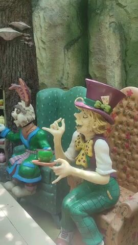 The video shows a scene with cartoon figures of the Mad Hatter and the March Hare from Alice in Wonderland sitting at a table laden with teacups and teapots. The Mad Hatter is on the right, sitting in a chair, dressed in green plaid pants, a vest, and a purple top hat with a "10/6" card on it. He's holding a teacup on a saucer and gesturing with his hands. The March Hare is seated to the left, wearing a green robe with a blue and pink sash. He appears to be reaching for a teapot on a shelf built into a tree trunk behind him. The table in front of them is covered with colorful teacups and teapots. The background suggests an indoor, artificially themed environment, with a faux rock wall and painted greenery visible.
