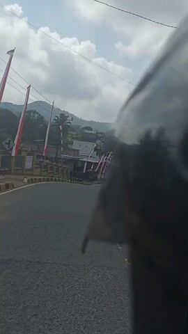 The video shows a view from a moving vehicle, likely a motorcycle, looking down a paved road lined with buildings. The road curves to the left and then straightens out, leading towards a distant hilly landscape under a partly cloudy sky. Along the sides of the road, there are numerous buildings, many of which appear to be shops or houses. Red and white flags, possibly national flags, are displayed prominently along the road, suggesting a festive or patriotic occasion. Several motorcycles are visible on the road, with people riding them. The overall scene depicts a street in a town or village, possibly in Southeast Asia, given the flags and architecture. The perspective suggests the video is being recorded by someone riding as a passenger or the driver themselves.