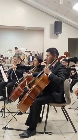 The video shows a section of an orchestra during a performance. Several musicians, mostly dressed in black suits and ties, are playing cellos. A few violinists are also visible. The camera pans slightly from left to right, revealing more of the cello section. The background includes other musicians, percussion instruments, and a banner that says "Sinfónica Del Cusco" in Spanish. The room is well-lit with white walls and a high ceiling. A black speaker is mounted on the wall. The overall atmosphere is formal and focused on the musical performance.
