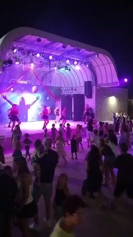 This video depicts an outdoor evening event with a stage in the background illuminated by purple and white lights. Several performers are on the stage, some appearing to be dancing. In front of the stage, a crowd of people, including adults and children, are gathered and also appear to be dancing. Some individuals are holding up their phones, possibly recording the event. The environment seems to be an open area, possibly a park or an entertainment venue, with some overhead structures and lights visible. The overall atmosphere appears lively and celebratory.