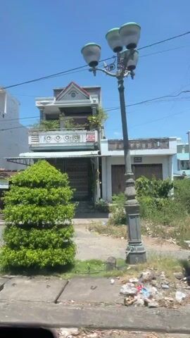 This video shows a daytime view from a moving vehicle traveling along a street in Đông Hòa, Vietnam. The scene is a mix of residential and commercial buildings lining the street, interspersed with lush greenery.

Dominant subjects include several multi-story buildings, some with balconies and decorative architectural elements. One building features a bright yellow and white facade. Another has a pale green and white exterior with a decorative balcony. Shops with various storefronts, including roll-up doors and display windows, are visible. A large, gray truck with a green trailer is seen driving on the road. There are also several trees and bushes, some of which are adorned with vibrant pink and orange flowers, creating a colorful contrast against the blue sky. A white car is parked in front of one of the shops.

The activity taking place is the vehicle's movement along the street, capturing the unfolding scenery. The time of day appears to be late morning or early afternoon, given the bright sunlight and clear blue sky, suggesting warm weather. The presence of shops, residential buildings, and vehicles indicates a typical urban or semi-urban environment in Vietnam. No people are clearly discernible as subjects within the frame.

Visible text includes signage on the buildings. One sign has yellow lettering on a black background that reads "TÍNH QUANG" with smaller text below it "LAPTOP - CAMERA". Another sign above a shop reads "Shop THẢO HIỀN" with smaller text below indicating "ÁO QUẦN NAM, NỮ HỢP THỜI TRANG". A sign on the side of the truck appears to have a logo and some Vietnamese text, but it is not fully legible. Another sign on a building in the background is partially visible with Vietnamese text. The overall mood conveyed is one of everyday life and activity in a Vietnamese town.