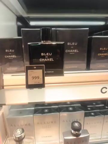 The video shows a retail display of Chanel perfumes. In the foreground, a bottle of "BLEU DE CHANEL EAU DE PARFUM" is displayed prominently. Next to it, a digital price tag shows "CHANEL" at the top, followed by product details and a price of "999 DKK" and "137.66 EUR". In the background, several boxed versions of the same perfume are visible, along with other Chanel products like "ALLURE HOMME SPORT". The lighting suggests an indoor retail environment, likely an airport duty-free shop, given the currency displayed.