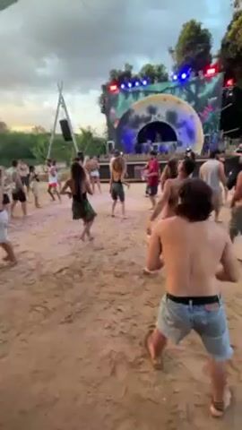 A crowd of people is dancing at an outdoor event. The ground is sandy and there are trees in the background. A stage with psychedelic decorations is visible. Colorful canopies hang overhead. Most people are dressed in casual summer attire, some are shirtless. Some people are standing around talking, while others are actively dancing. The sky is cloudy, suggesting it might be sunset.