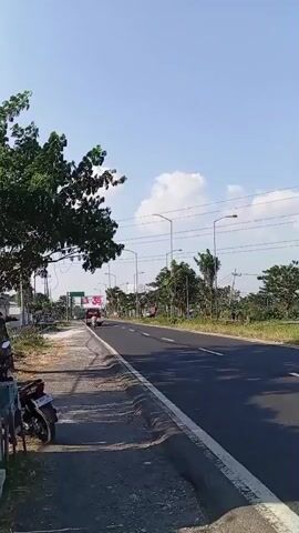 A vertical video shows a two-lane road with a clear blue sky and some clouds. On the left side of the road, there is a dirt shoulder with a tree, a few parked motorcycles, and a small building. On the right side of the road, there is a paved shoulder, streetlights, and trees lining the road. Traffic is moving in both directions. Several motorcycles and a few cars are visible on the road. A large billboard with the number "35" and some text is visible in the distance. The video appears to be filmed from a moving vehicle.