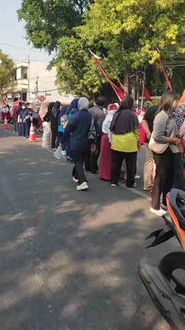 The video shows a street scene with people lining up along the side of the road. Many people are wearing school uniforms, consisting of white shirts and red or green skirts or pants, along with headscarves for some. There are also adults in casual clothing, some appearing to be supervising or accompanying the students. Orange traffic cones are placed along the line to demarcate the area. Trees line the opposite side of the street, and buildings are visible in the background. The sky is clear, suggesting daylight. Some individuals are holding plastic bags. The overall impression is of an organized gathering, possibly a school event or parade.