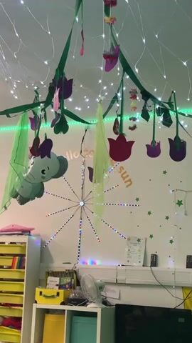 This video captures a brightly decorated classroom, likely in Wakefield, United Kingdom. The ceiling is adorned with a whimsical display of hanging greenery, colorful cut-out tulips and other floral shapes, and an abundance of twinkling fairy lights. A large inflatable elephant hangs from the ceiling, adding a playful element.

A central feature on the wall is a starburst-shaped light fixture with individual colored LEDs. Above this, the words "Hello to the sun" are visible in large, stylized lettering. The walls are decorated with green star cut-outs, creating a starry night or whimsical garden theme.

On the shelves below, various objects are visible, including storage bins, a fan, and what appear to be educational materials. The overall impression is one of a cheerful and stimulating environment, possibly set up for a special event or a themed lesson. The lighting, with its mix of white fairy lights and the colorful starburst, suggests the activity might be taking place indoors during daylight or at night, as the lights create a festive and magical atmosphere. There are no people prominently featured in the video, but the presence of classroom furniture implies an educational setting.