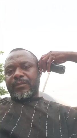 A man is getting a haircut outdoors during the daytime. The client, a dark-skinned man with a beard (which has some grey hairs), is wearing a dark brown or black shirt with thin white vertical stripes. He is looking directly at the camera with a calm expression.

A barber, a dark-skinned man wearing a black and red striped/plaid shirt, is standing behind him and is in the process of shaving the client's sideburn and around his ear using an electric hair clipper. The barber's hand is holding the clipper, and his other hand is holding a black cloth near the client's ear.

The background consists primarily of a bright, clear or slightly overcast sky, indicating an outdoor setting. Some green foliage is visible on the far left of the frame. The overall impression is that of a casual, outdoor haircut.