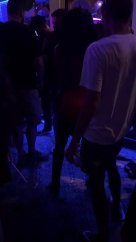 The scene is a dark, crowded nightclub with blue ambient lighting. Several people are present, including a man in a white t-shirt and dark shorts, dancing to Latin music. He is wearing flip-flops. Other people are indistinctly visible in the background, some leaning against a bar with tropical decor. One person is wearing cowboy boots. The floor appears to be covered in a pebble-like material or gravel. The lighting shifts occasionally with strobes.