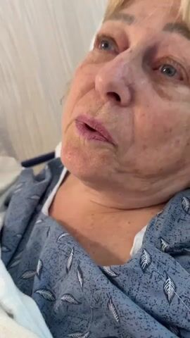 The video shows a close-up of an elderly Caucasian woman with blonde hair and blue eyes. She is wearing a blue and white patterned hospital gown and is lying down, likely in a hospital bed, as indicated by the white bedding and a blue pole visible in the background. Her facial expressions suggest she is speaking or reacting to something, with her mouth slightly open and her eyes looking upwards or to the side. Her skin has some age spots and redness, and her lips are lightly colored. The lighting appears to be from above and slightly to the right.