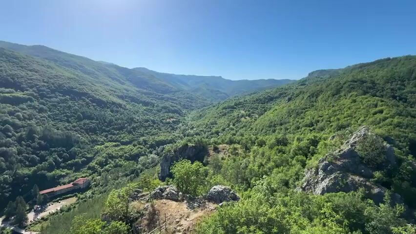 This is a video of a vast green mountain landscape under a clear blue sky. The camera pans from left to right, revealing rolling hills covered in dense trees. In the foreground, there are rocky outcrops and some greenery. In the midground, a valley can be seen with some scattered buildings and more trees. The sun is shining brightly, casting long shadows and highlighting the textures of the landscape. The overall impression is one of natural beauty and tranquility.