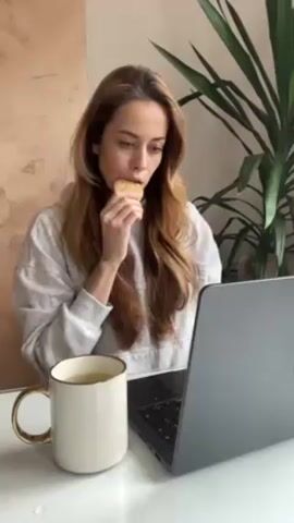 A woman with long brown hair, wearing a gray hoodie, is sitting at a white desk in front of a gray laptop. She is eating a round cookie and looking at the laptop screen. To the left of the laptop is a white mug with a gold handle. Behind her is a painted wall and a large green potted plant. There is a drop of liquid on the desk.