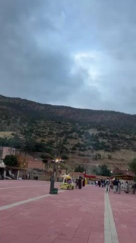 The video shows a wide-angle view of a town square or plaza with a mountain in the background under an overcast sky. The ground is mostly paved with red tiles and has white stripes. Several people are walking around, some in traditional clothing. There are a few small vendor stalls and what looks like a small electric amusement ride shaped like a car. A tall light post stands in the foreground. The mountain behind is covered with sparse trees and vegetation. The town buildings are visible on the left side of the frame. There is no sound in this short video.